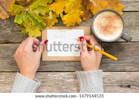 Autumn still life. Woman's hands holding a pen writing Hello autunm text on paper with craft envelope. Cup of coffee, cappuccino, latte, autumn leaves aside on vintage background. Hot drink for