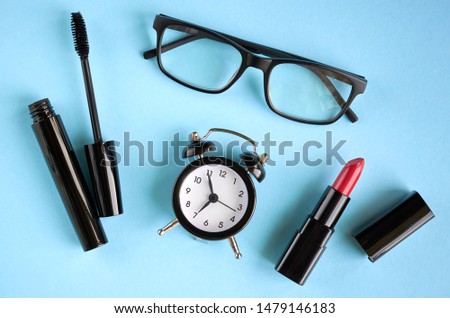 Black glasses, alarm clock, mascara and red pomade on blue background composition. Flat lay and top view photo