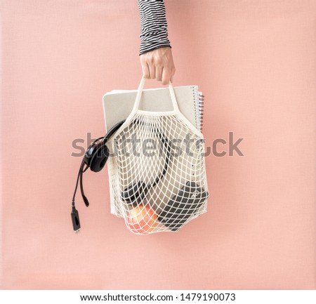 Hand holding mesh shopping bag with notebooks, headset, apple and smartphone. Back to school, education concept. Canvas background.