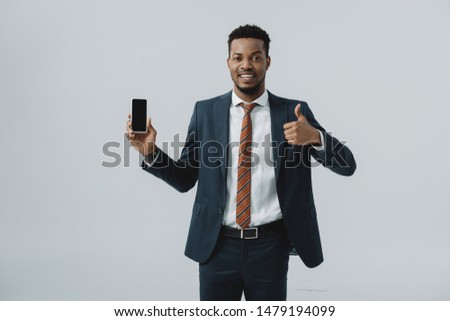 happy african american businessman showing thumb up and holding smartphone with blank screen isolated on grey