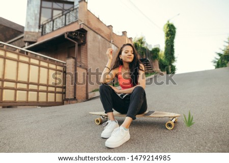 Smiling  girl sitting on logboard on street background, looking down, wearing street clothes. Girl skater relaxing on longboards on asphalt, positive, street culture.
