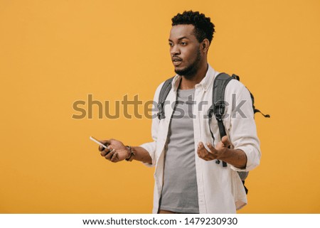 emotional african american man with backpack holding smartphone and gesturing isolated on orange