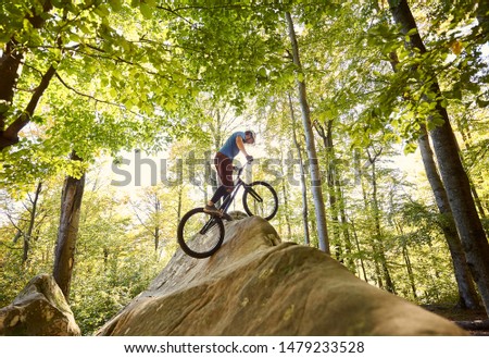 Athlete male cyclist balancing on trial bicycle, biker making acrobatic stunt on big boulder in the forest outdoor on summer sunny day. Concept of extreme dangerous sport active lifestyle