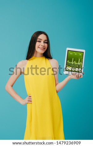 smiling brunette girl in yellow dress showing digital tablet with healthcare app isolated on turquoise