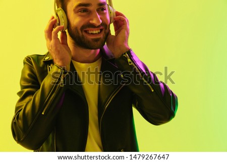 Picture of smiling pleased handsome young stylish man posing isolated over light green background wall with led neon lights dressed in leather jacket listening music with headphones.