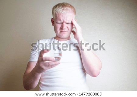 A man with blond hair in a white T-shirt suffers from a headache. In a hand is a glass of water. On the face is an expression of pain and fatigue. The concept of pain medication, migraine, hangover.