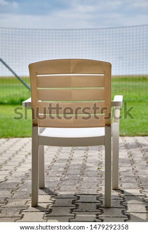 An empty white garden chair is faced out on some fields, meadows and a high fence on a sunny summer day