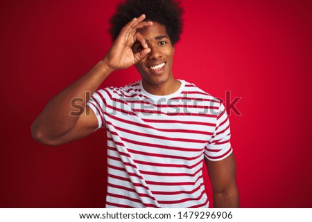 Young african american man with afro hair wearing striped t-shirt over isolated red background doing ok gesture with hand smiling, eye looking through fingers with happy face.