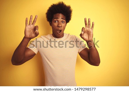 American man with afro hair wearing striped t-shirt standing over isolated yellow background looking surprised and shocked doing ok approval symbol with fingers. Crazy expression