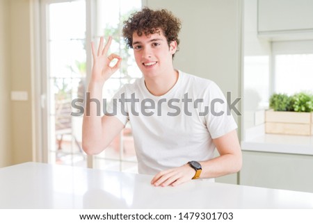 Young handsome man wearing white t-shirt at home smiling positive doing ok sign with hand and fingers. Successful expression.