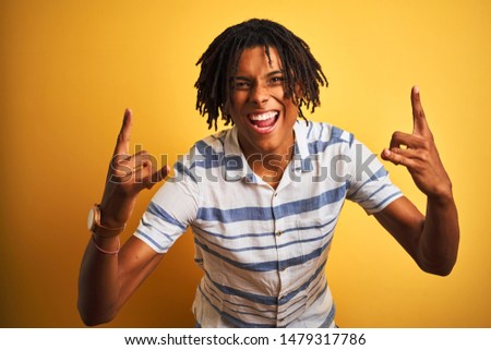 Afro american man with dreadlocks wearing striped shirt over isolated yellow background shouting with crazy expression doing rock symbol with hands up. Music star. Heavy concept.