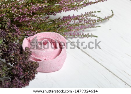 Pink marshmallows in the form of a flower in icing sugar. Lies on white painted boards. Nearby lies a bouquet of dried thyme and fresh heather.