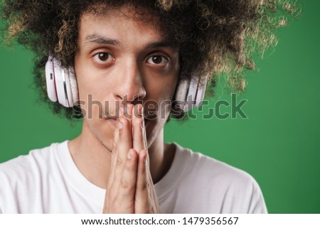 Photo of a young serious curly guy posing isolated over green wall background listening music with headphones make hopeful please gesture.