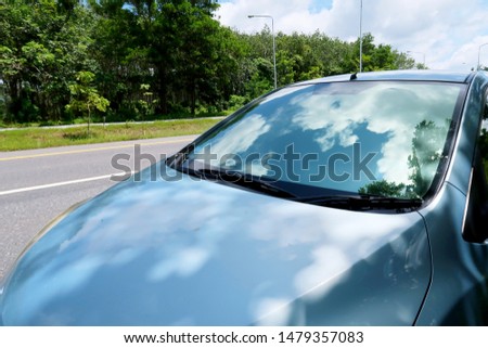 Front view of blue shiny empty car with Reflection of white clouds blue sky on mirror in summer bright sunny day beside asphalt road near  green rubber plantation forest, copy space                  Royalty-Free Stock Photo #1479357083