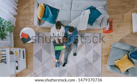 Young Couple is Sitting on a Floor and Using a Laptop with Green Screen. Girl Drinks Coffee. Cozy Living Room with Modern Interior with Carpet, Sofa, Table, Shelf, Plant and Wooden Floor. Top View.
