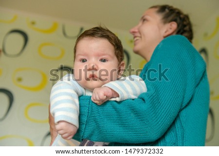 In the arms of a cheerful mom. Portrait of charming toddler with blue eyes and chubby cheeks resting vertically in his mother's arms. Beautiful young European mom with loose ginger hair bonding with