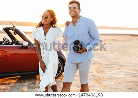 Photo of beautiful multiethnic couple man and woman hugging together and walking with retro camera by car outdoors