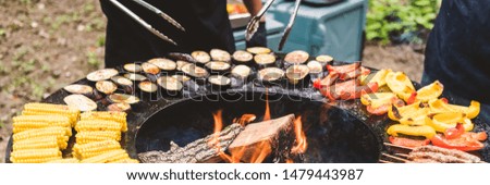 Round barbecue grill with open fire inside. Meals for the summer picnic are being prepared: corn, eggplant, bell pepper, kebab. Male hands in black gloves turn the food over with barbecue tongs