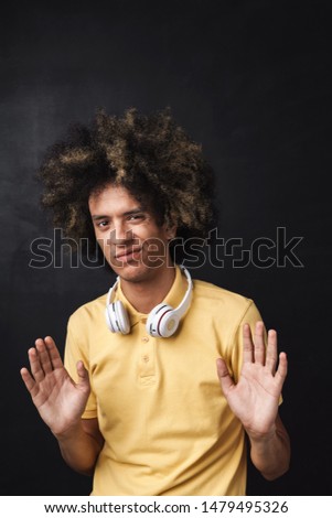 Photo of a curly young displeased teenage guy posing over grey chalkboard wall with headphones showing stop gesture.