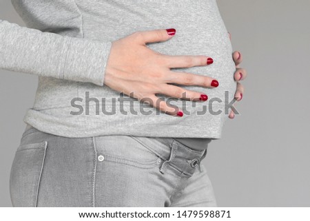 Closeup of torso and hand of young pregnant model standing and caressing her belly, isolated on the grey background. Future mom expecting baby. Maternity concept.