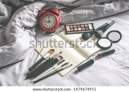 Make up cosmetic and brushes, alarm clock and blank page notepad on the blanket on the bed background. Female morning makeup tips mockup.