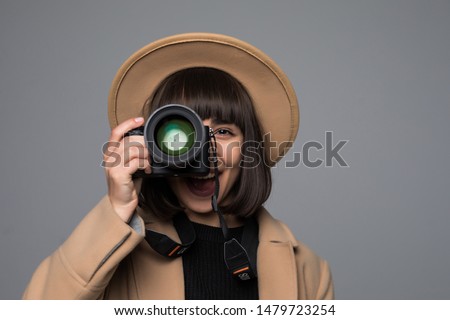 Portrait of young woman wears hat and beige coat takes pictures with her camera isolated on gray background.