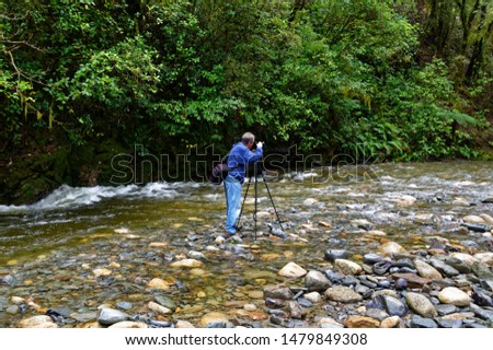 A male photographer is standing in the middle of a river with his camera on a tripod taking a photo