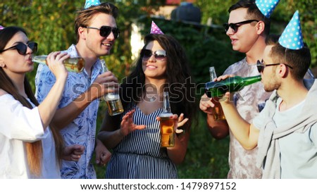 Group of young people drinking beer from bottle, friends dancing on summer party, wearing birthday caps
