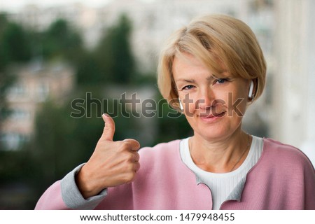 Portrait of cheerful elderly adult woman listening music in wireless earphones, show thumb up. Senior mature female pensioner smiling outside. Happy retirement. Good looking old person and technology.