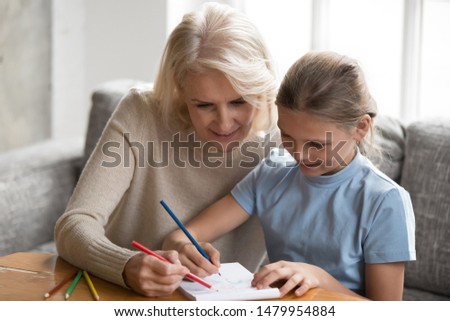 Grandmother spend time at home with granddaughter sit on sofa drawing with colored pencils in album, retired nanny teach little girl help create picture, leisure activity different generations people