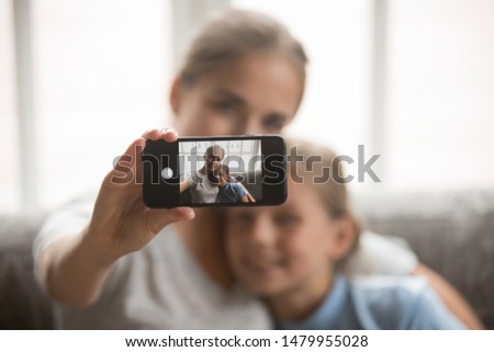 Young beautiful mother and little daughter sit on couch at home embracing making selfie view through mobile screen, younger and older sisters taking photo using modern wireless gadget concept image