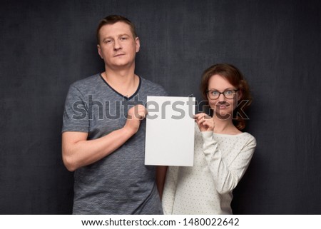 Studio half-length portrait of caucasian couple dressed casually, holding white blank paper sheet together with place for your text, being calm and positive, standing over gray background