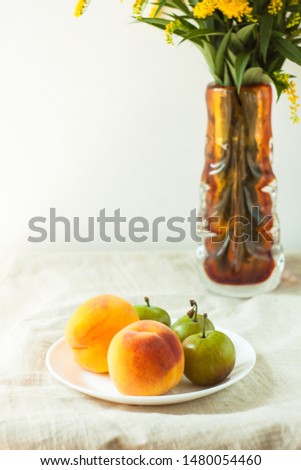 Still life with fresh peaches and plums on linen cloth background on wooden table. Bright juicy summer fruits on a white plate with yellow flowers in a vase.Breakfast,healthy raw vegan food,front view
