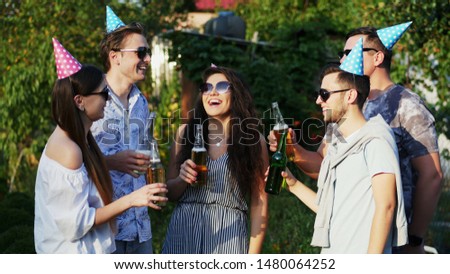 Group of happy friends clink with beer bottles on summer party while celebrate birthday, say toasts, people in holiday caps