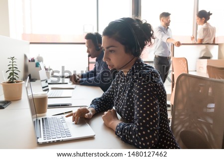 Attentive call centre indian woman employee sitting in modern shared office working using computer wearing headset with microphone answering or making telephone calls, give support to company clients Royalty-Free Stock Photo #1480127462