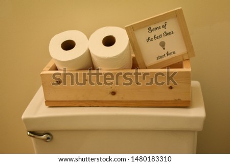 some of the best ideas start here sign in wooden frame tucked into toilet paper wooden box on white toilet with flusher tan background wall