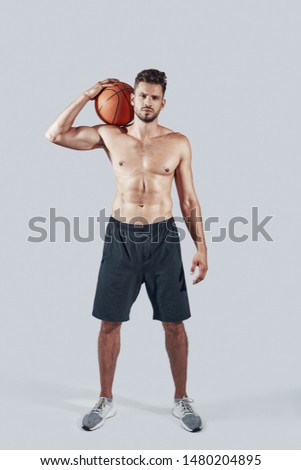 Full length of handsome young man in sports clothing carrying basketball ball and looking at camera while standing against grey background