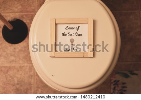 sign that reads some of the best ideas start here on a white toilet seat bathroom floor background black and wood plunger and corner of flowered bathroom rug