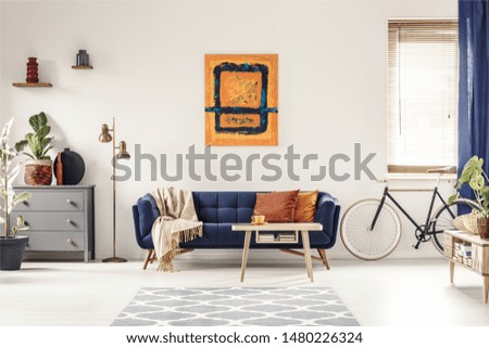 Yellow and blue painting hanging on white wall in bright living room interior with grey cupboard, gold lamp, sofa with blanket and pillows, and bike standing under window with blinds Royalty-Free Stock Photo #1480226324