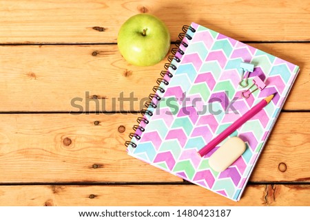 Top view of girly pink notebook with pencil, eraser, binder clips and apple on wooden background.