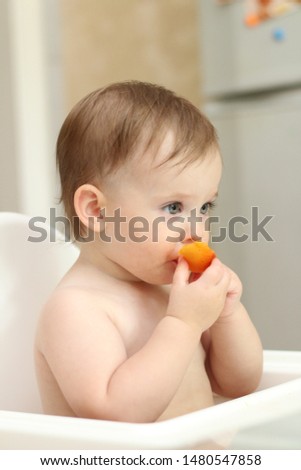 cute blond child sits in a children's chair at the kitchen and eat a juicy peach. Little girl with a piercing look