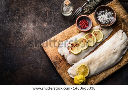 Raw cod fish fillet with lemon slices and herbal salt on rustic wooden cutting board on dark background, top view. Fish cooking preparation. Healthy diet food. Border. Copy space
