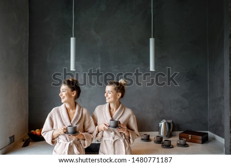 Two young women in bathrobes standing together on the kitchen of the modern SPA complex or home, drinking coffee after the SPA procedures