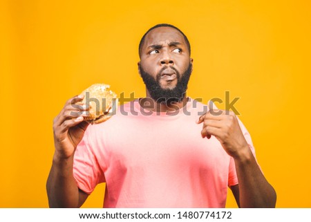 Young african american man eating hamburger isolated over yellow background.