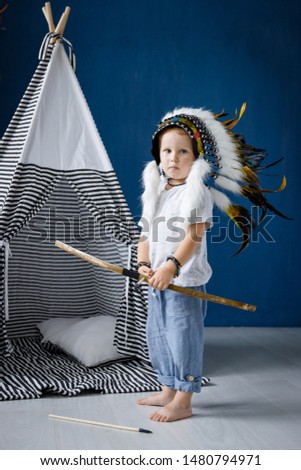 A little boy plays Indians with a bow and arrow in a big feather Mohawk next to a wigwam.