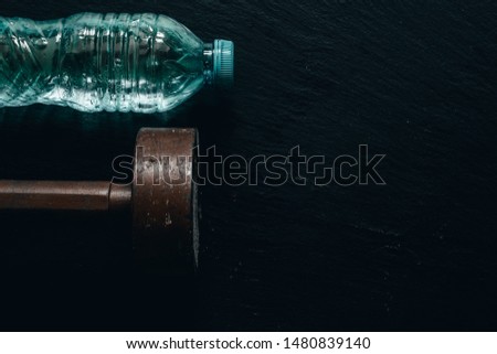 Heavy brown dumbbell with water bottle on black background with copy space - Old and rusty gym weight used for bodybuilding with plastic liquid container on dark stone textured surface