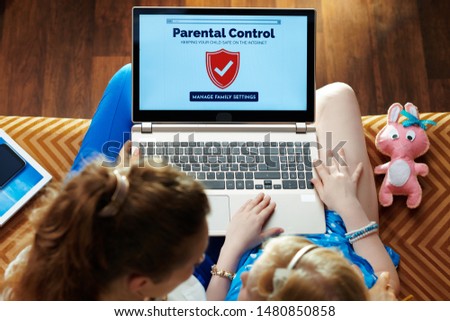 modern mother and daughter sitting on sofa in the modern living room setting up parental control on a laptop.