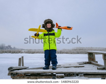 Active funny kid in bright clothers play on snowy river. Boy launch bright toy planes. Christmas Eve. Winter holidays in village. Family holidays.