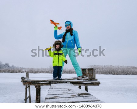 Slender cheerful mother in blue ski suit plays with children on snowy river. Boys with mom launch bright toy planes. Christmas Eve. Winter holidays in village. Family holidays.