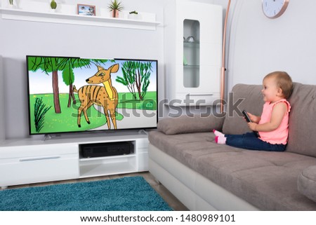 Little Baby Girl Sitting On Sofa Watching Cartoon On Television At Home